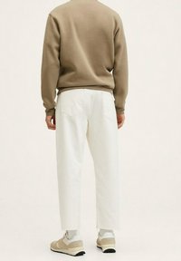 Persona in piedi di spalle, indossa una felpa beige, pantaloni bianchi larghi, calzini bianchi e sneakers beige su sfondo semplice.