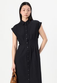 Vestido preto de camisa com mangas curtas enroladas, frente com botões, cintura pregueada e saia. Modelo segura uma pequena bolsa castanha.