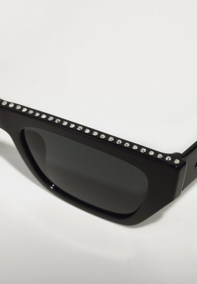 Occhiali da sole neri con lenti scure, caratterizzati da una fila di strass trasparenti lungo il bordo superiore della montatura. Design quadrato elegante con finitura opaca.
