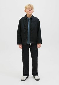 Blondt barn står vänd framåt iförd en svart zip-up jacka, blå skjorta, svarta cargobyxor och vita sneakers med svarta accenter.