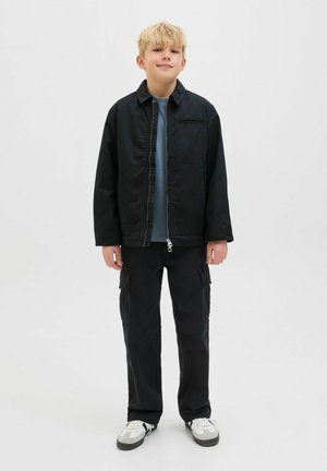 Un enfant blond se tient face à l'avant, portant une veste zippée noire, une chemise bleue, un pantalon cargo noir et des baskets blanches avec des accents noirs.