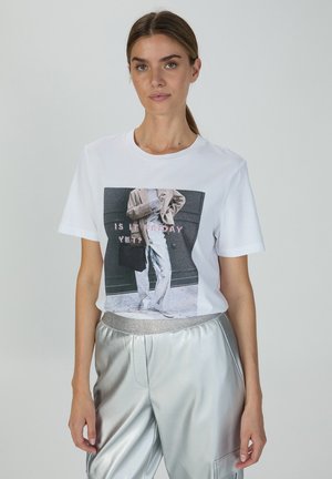 Weißes Baumwoll-T-Shirt mit grafischem Druck auf der Vorderseite und Text. Kombiniert mit glänzenden silbernen Metallic-Hosen. Kurze Ärmel, runder Ausschnitt.