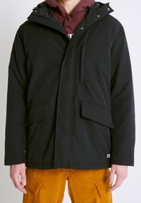 Chaqueta aislante negra con capucha, que presenta un bolsillo en el pecho con cremallera, dos grandes bolsillos frontales y un acabado texturizado.