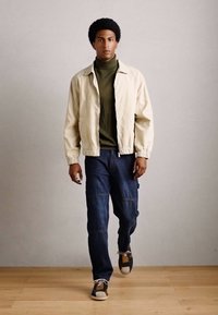 Young man walking indoors wearing beige jacket, olive green turtleneck, dark blue jeans, and black-and-white sneakers on wooden floor.
