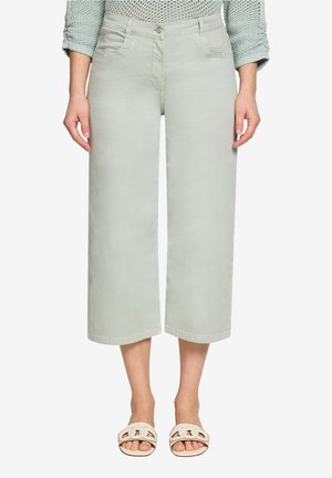 Femme portant un pantalon large court gris clair, un haut en maille vert menthe et des sandales blanches à bout ouvert sur un fond uni.