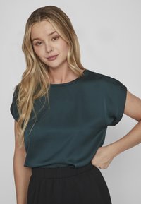 Mörk teal, kortärmad blus med en slät, glänsande textur och rund halsringning. Bärs med en svart, veckad kjol.