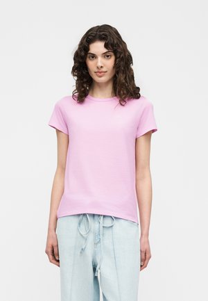 Młoda kobieta z kręconymi ciemnymi włosami, ubrana w jasnoróżowy t-shirt z krótkim rękawem i jasnoniebieskie spodnie ze sznurkiem, stoi na jednolitym tle.