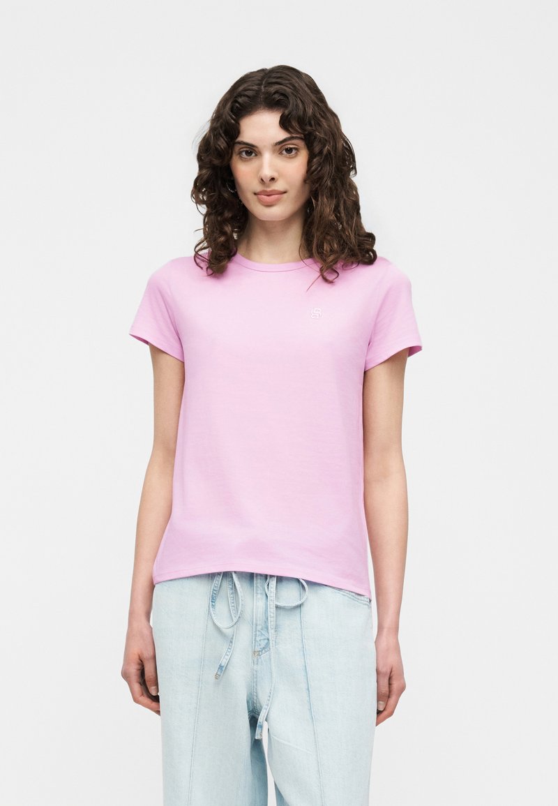 Νέα γυναίκα με σγουρά σκουρόχρωμα μαλλιά που φοράει ένα ανοιχτό ροζ κοντομάνικο μπλουζάκι και ανοιχτό μπλε παντελόνι με κορδόνι, στέκεται μπροστά από φόντο μονής απόχρωσης.