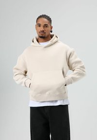 Beige hoodie met een zak aan de voorkant, capuchon met trekkoord en geribbelde manchetten. Draagt hem over een wit T-shirt en combineert met een zwarte spijkerbroek.