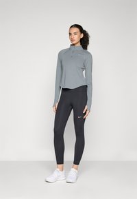 Maglia atletica a maniche lunghe in grigio chiaro con zip quarti abbinata a leggings scuri. Entrambi presentano una vestibilità slim e dettagli con logo. Scarpe sportive bianche completano il look.