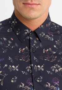 Button-up shirt in dark navy fabric, featuring a floral pattern in various colors. Notable details include a classic collar and black buttons.