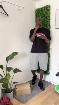 Black t-shirt with logo, gray shorts, black sneakers. Indoor setting with potted plant, green wall accent, and cardboard box on a rug.