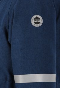 Marineblaue Jacke aus einem strukturierten Stoff; mit einem kreisförmigen Zickzack-Logo auf dem Oberarm und einem reflektierenden silbernen Streifen.