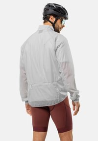 Lichte, ademende grijze fietjas met lange mouwen, een achterzak met rits en een geventileerd ontwerp. Draagt over bordeauxrode fietsshorts.