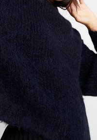 Osoba poprawiająca ciemnonawizowy, puszysty, dziany sweter o grubym splocie, nosząca czarną spódnicę na jednolitym tle.