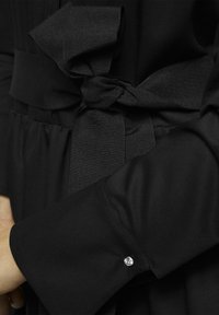 Schwarzes Kleid mit einem strukturierten Bandgürtel, der auf Taillenhöhe gebunden ist. Die Ärmel haben einen Bund, der mit einem kleinen dekorativen Stein verziert ist. Glattes Material.
