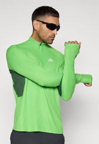 Πράσινο αθλητικό τοπ με μακριά μανίκια και φερμουάρ μέχρι το στήθος, με υφή υφάσματος και σκούρες πράσινες πλαϊνές επενδύσεις. Αντανακλαστικό λογότυπο στο στήθος.