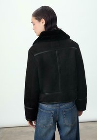 Czarna kurtka z owczej wełny z szerokim kołnierzem, z teksturowanej tkaniny i kontrastującymi skórzanymi akcentami. Model ma na sobie luźne niebieskie dżinsy.