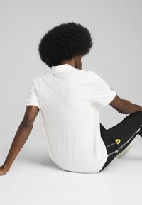 Hvid polo shirt med korte ærmer, der har en krave og side-striber. Kombinationen er med sorte bukser, der viser et logo og hvide detaljer.