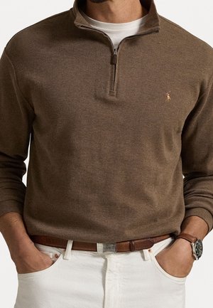 Man wearing brown quarter-zip sweater over white shirt, white pants with brown belt, wristwatch on left wrist, hands in pockets.