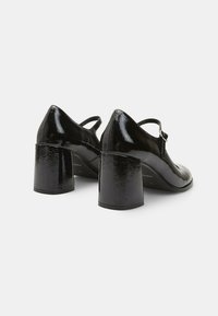 Black patent leather heels with a square toe, curved design, closed back, and chunky block heels, featuring an adjustable strap detail.