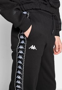 Zwarte sportieve joggers met een trekkoord in de taille, voorzien van grijze Kappa-logo's langs de zijkant en een zachte textuur. Inclusief zijzakken.