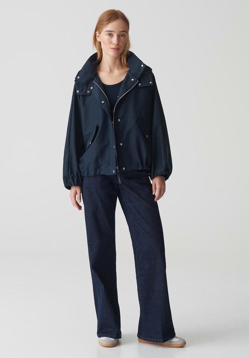 Donkerblauwe lichtgewicht jas met een grote capuchon, ritssluiting aan de voorkant en elastische manchetten, gecombineerd met donkere denim wijde broek en lichte schoenen.