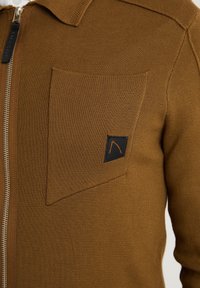 Hnědá pletená zipová mikina s límečkem a kapsou na levé straně hrudi, která má černý nášivku a oranžový logotyp.