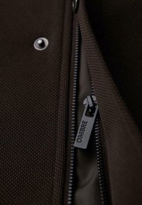 Tela marrón con una superficie texturizada, que presenta una cremallera negra con la marca "OMBRA" y un detalle de botón de presión al lado de la apertura.
