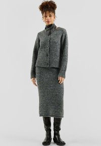 Frau in einer grauen Strickjacke und passendem Midirock, schwarzen Lederstiefeln und einem schwarzen Halstuch, stehend vor einem einfarbigen Hintergrund.