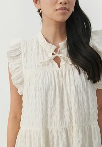 Robe blanche texturée avec des manches volantées, une ouverture en trou de serrure et un détail noué au niveau du col. Tissu doux et coupe décontractée.