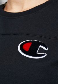 Černá mikina s kulatým výstřihem, s texturovaným vyšitým logem v červené a bílé barvě na levé straně.