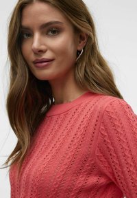 Koralstrikket sweater med et tekstureret mønster, ribbet halsåbning og korte ærmer. Modellen har bølget hår og bærer sølvfarvede hoops.