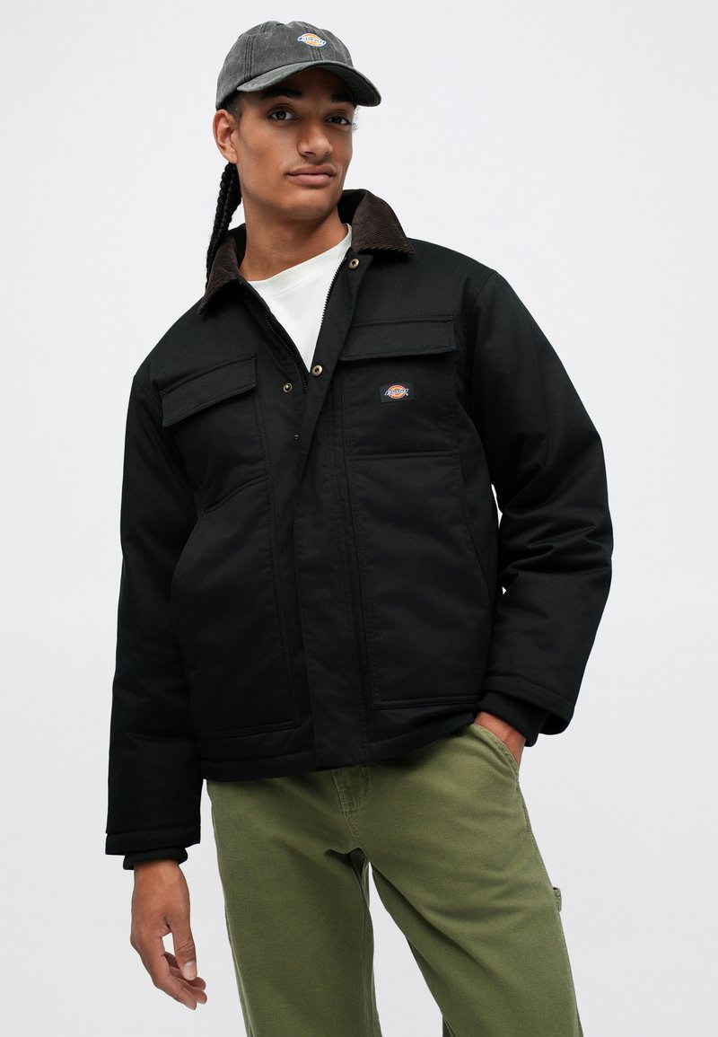 Giovane con lunghi capelli intrecciati, indossa una giacca nera con colletto marrone, pantaloni verdi, berretto grigio e camicia bianca su uno sfondo neutro.