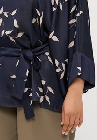 Blusa blu navy con un motivo floreale in beige chiaro, con maniche ampie, vita con fiocco e texture liscia in satin.