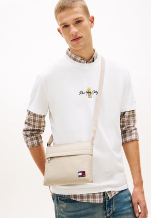 Bolso bandolera beige hecho de tela texturizada con cierre de cremallera, que incluye un parche con el logo. El modelo lleva una camisa blanca sobre una camisa de franela de cuadros.