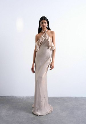 Mujer de cabello largo y oscuro viste un vestido beige largo hasta el suelo con cuello halter y volantes, de pie sobre un suelo de concreto gris frente a una pared blanca y lisa.