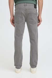 Mees, kes kannab hallid sirgelõikelisi teksapükse ja valgeid vabaajajalatsid, seisab valge tausta ees, käed lõdvalt külgedel.