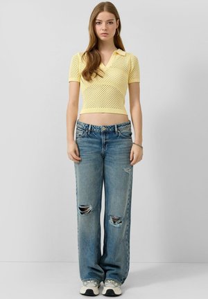 Junge Frau mit langen Haaren, die ein gelbes kurzärmliges Stricktop, zerrissene blaue Jeans und weiße Sneaker trägt und vor einem schlichten Hintergrund steht.