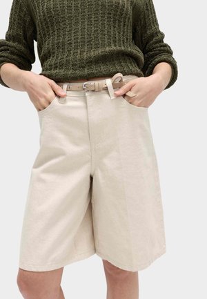 Person iført lysebeige højtaljede shorts med beige bælte, kombineret med en mørkegrøn tekstureret strikket sweater, hænderne i lommerne.