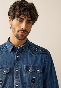 Camisa de mezclilla azul con dos bolsillos frontales, botones a presión blancos, costuras decorativas y acentos geométricos en los hombros.