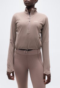 Γυναίκα που φοράει μπεζ μακρυμάνικο τοπ με φερμουάρ και αντίστοιχο κολάν με ψηλή μέση, στέκεται μπροστά σε απλό ανοιχτό φόντο.