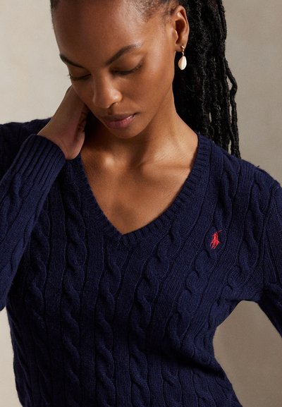 Woman with dark braided hair in a navy cable knit V-neck sweater with red logo, wearing a white shell earring, hand resting on neck.