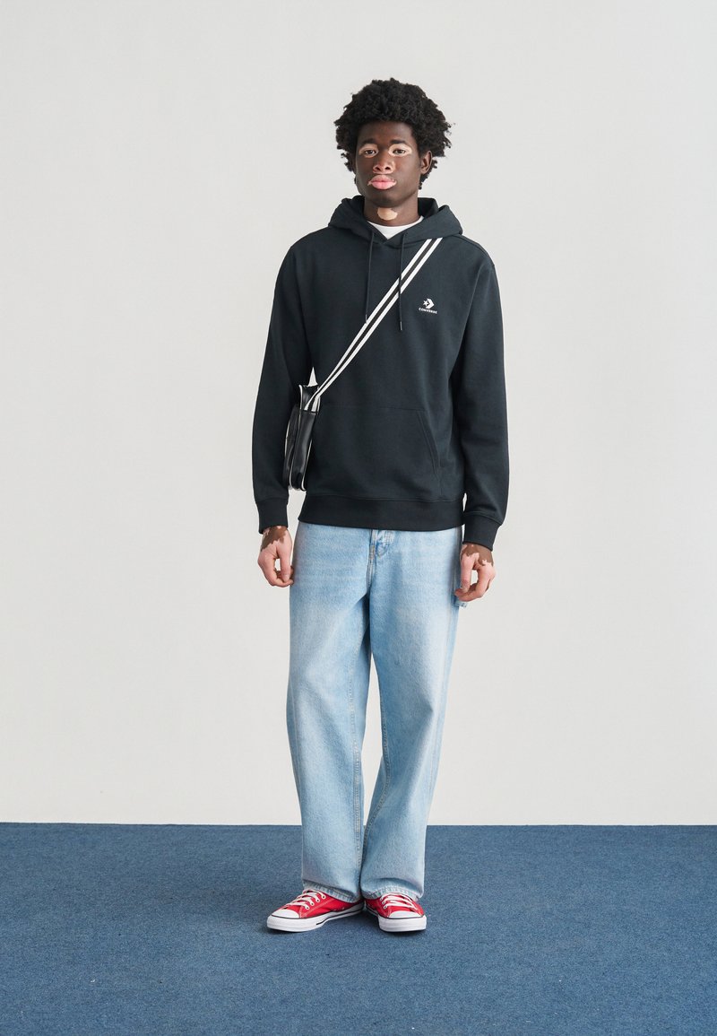 Sweat à capuche noir avec poche avant, jean bleu clair ample et baskets rouges. Le mannequin porte un sac bandoulière noir avec une bande blanche.