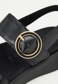Black leather sandal with a round gold-tone buckle detail, adjustable strap, cushioned black sole, and minimalistic design.