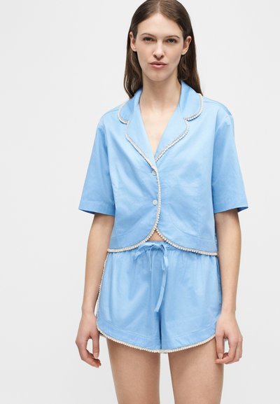 Femme portant une chemise à manches courtes bleue clair avec boutons et un short assorti à cordon de serrage avec une bordure blanche, debout devant un fond uni.