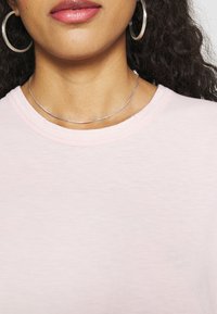 Light pink t-shirt with a round neckline, short sleeves, and soft fabric texture. Two silver necklaces and large hoop earrings are visible.