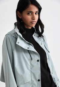 Light gray hooded jacket with a zipper and snap buttons, featuring a relaxed fit and drawstring details. Worn over a black sweater.