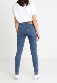 Vaqueros de denim azul ajustados con una cintura de altura media, bolsillos traseros y una etiqueta de cuero marrón. Llevados con una camiseta blanca y zapatillas blancas.