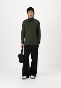Groene coltrui, losjes gesneden, gecombineerd met brede zwarte broek. Zwarte schoudertas met een schouderband, grijze en witte schoenen. Eenvoudige achtergrond.
